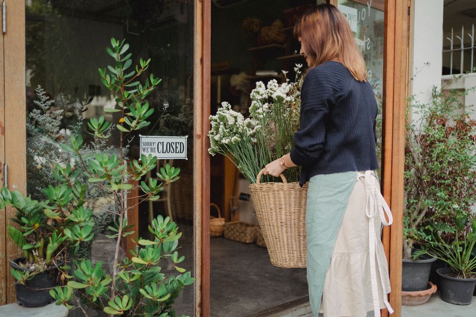 A woman with shoulder-length auburn hair, wearing a navy blue sweater and a light beige apron with a tied bow at the waist, stands at the entrance of a flower shop holding a large wicker basket filled