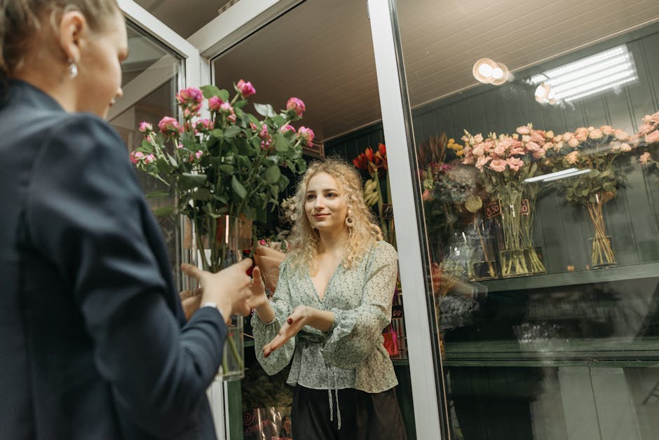 A floral arrangement is being handed over by a person wearing a dark jacket to a young woman with curly blonde hair and light skin inside a flower shop. The bouquet consists of pink roses with lush gr