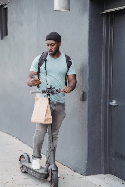 A young man with dark skin wearing a black beanie, light blue T-shirt, and grey jeans, standing next to a grey wall, holding a brown paper bag in one hand and a smartphone in the other. He is standing
