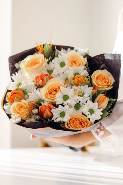 A person holding a bouquet of fresh flowers, featuring prominent peach roses with soft, velvety petals and white daisies with bright green centers. The arrangement includes small orange and yellow acc