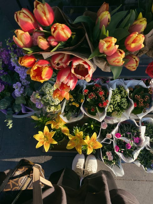 A vibrant display of fresh flowers arranged in bouquets and potted plants on a dark outdoor surface, viewed from a top-down perspective. The prominent bouquet features tulips with orange and yellow pe