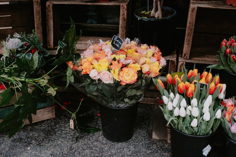 A black floral arrangement bucket filled with a vibrant bouquet of peach, pink, and yellow roses with lush green leaves, placed on a textured surface in a flower shop. To the right, a similar black bu