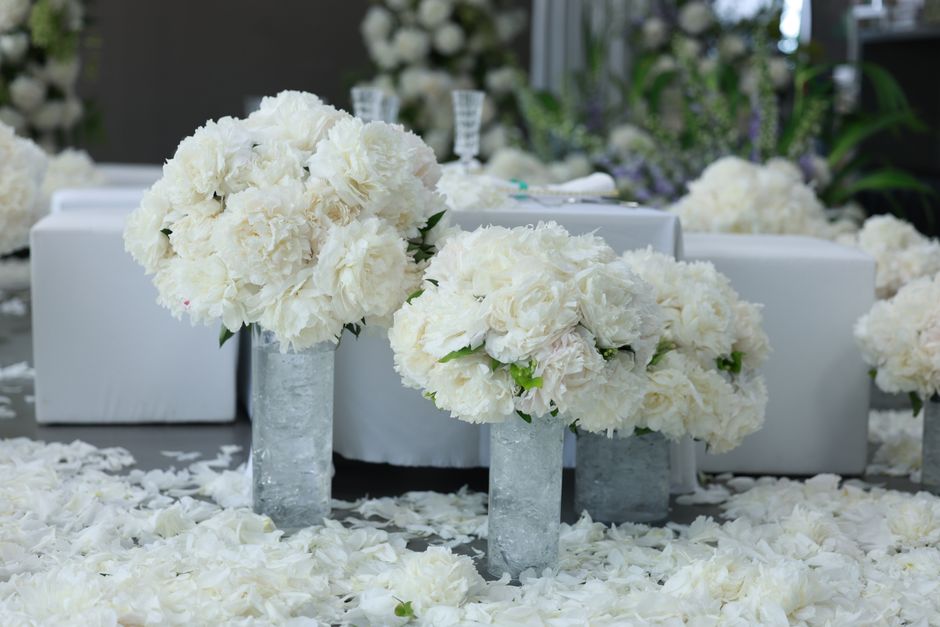 Two tall cylindrical vases filled with large, lush white hydrangea blooms and white roses, arranged as floral bouquets. The flowers are fresh, with delicate petals and green foliage peeking through. T