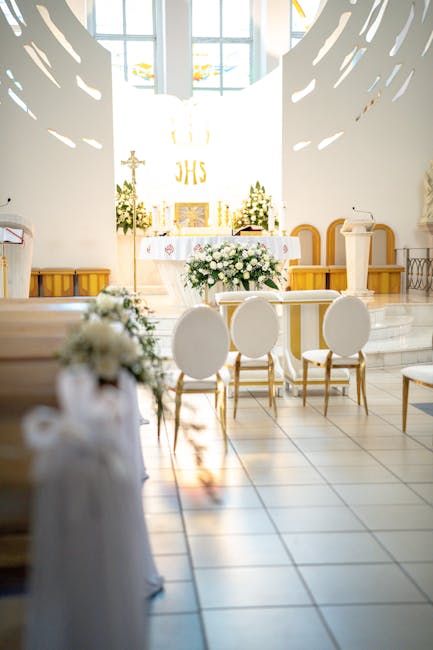 A softly lit church interior prepared for a wedding ceremony, featuring an altar decorated with a large, lush bouquet of white roses, lilies, and greenery. In front of the altar, there are six elegant